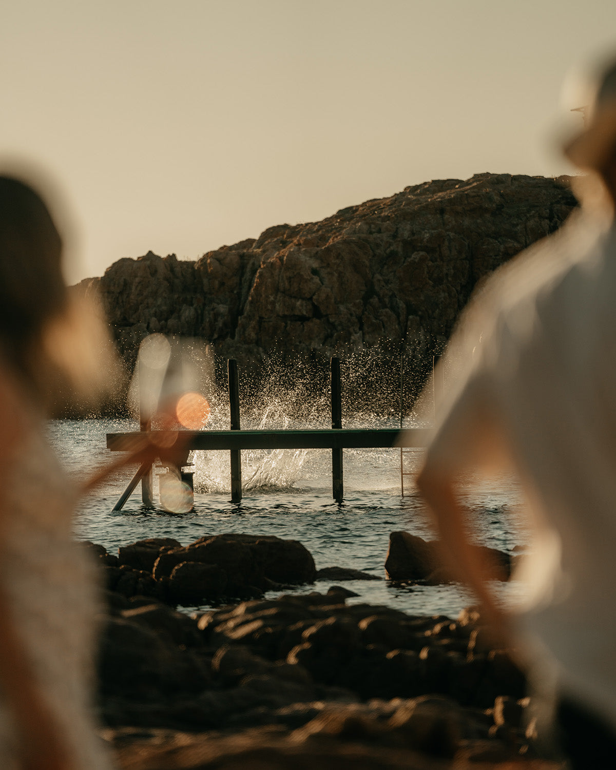 Two people standing on a rocky shore with a dock and water in the background