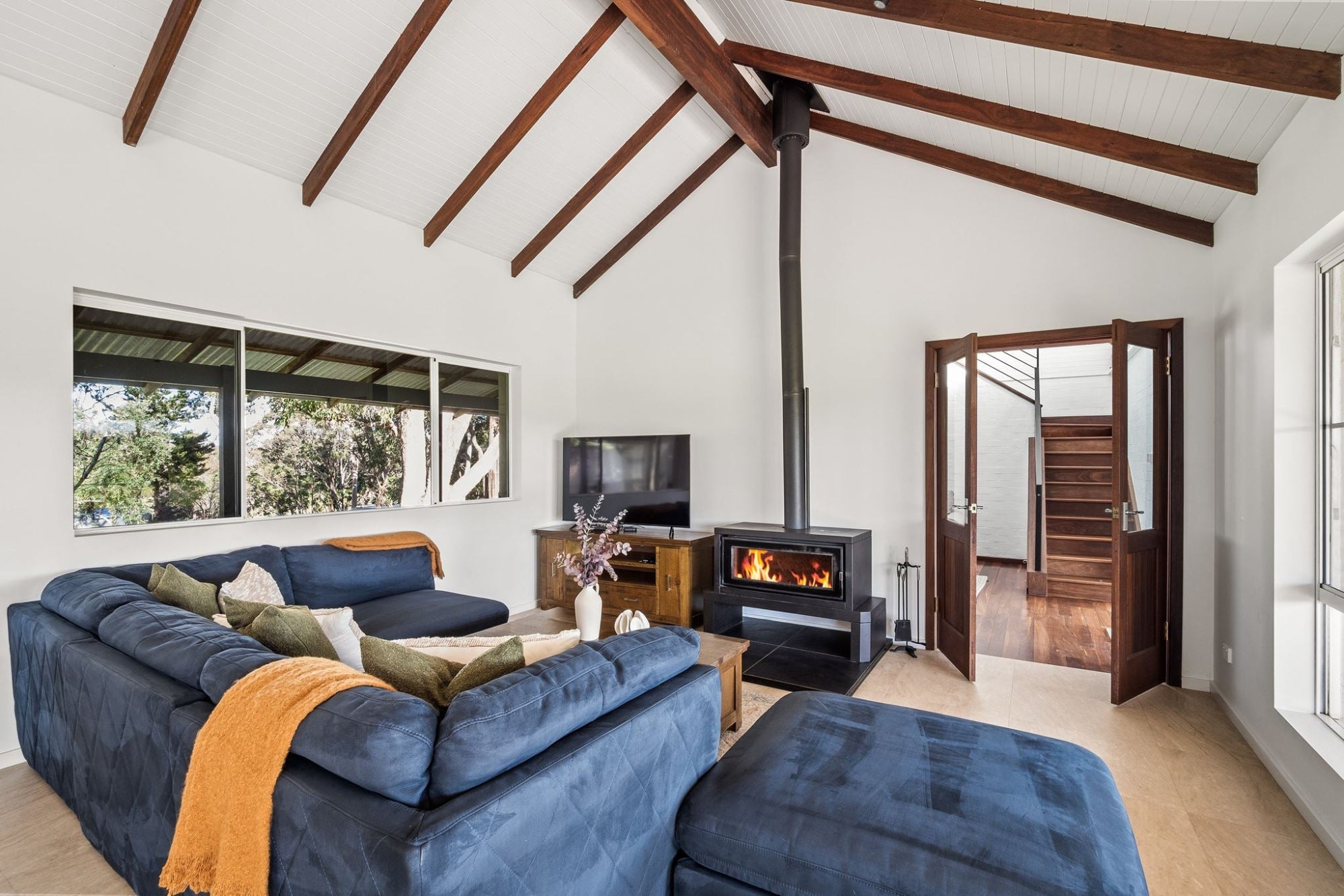 A modern living room with a large blue sectional sofa and matching ottoman, a black wood-burning stove with visible flames, exposed wooden ceiling beams, and an open doorway leading to a staircase.